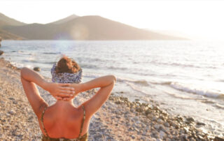 woman standing on a beach, with hands behind head, looking out at the ocean